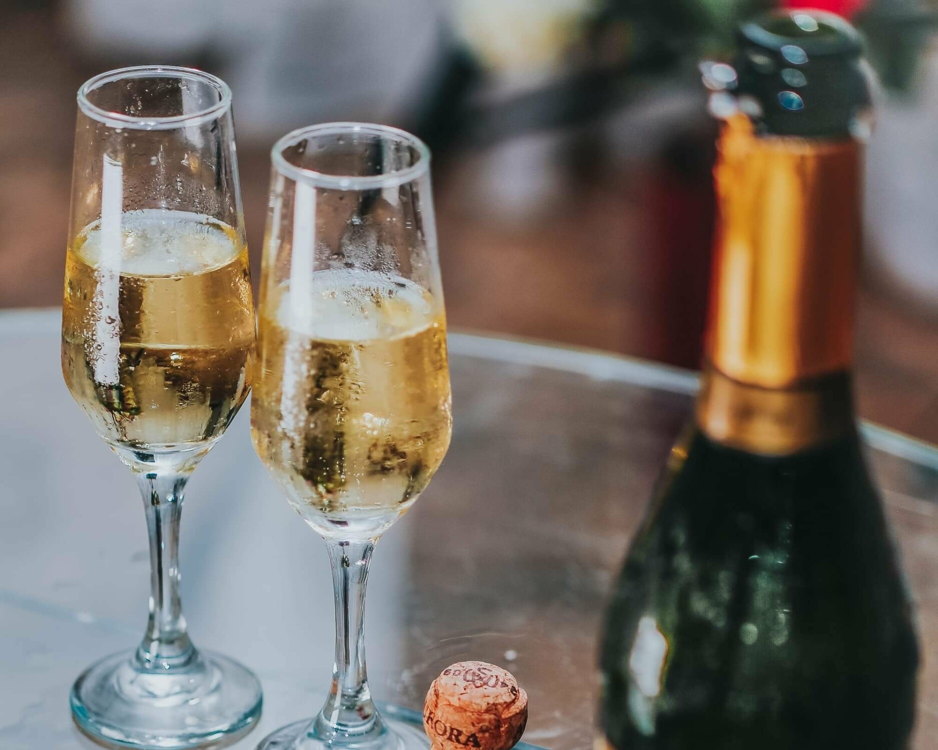 A picture of two glasses of champagne on a glass table next to a bottle of champagne and a cork to the right